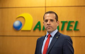 Vinicius Caram posa à frente de parede de madeira com o logo da Anatel ao fundo